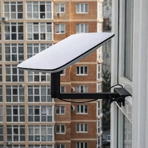 May include: A white rectangular satellite dish mounted on a black bracket, attached to a window frame. The dish is angled towards the sky, with a multi-story building in the background.