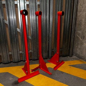 May include: Three bright orange metal stands with black circular tops and triangular bases are positioned on a grey concrete surface with yellow and black diagonal stripes. The stands are evenly spaced and appear unused.
