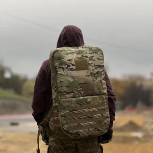 Puede incluir: Una persona lleva una mochila de camuflaje y pantalones a juego. La mochila tiene un bolsillo grande en la parte delantera y está hecha de un material duradero.