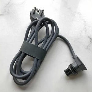 Op de afbeelding: Een grijs elektrisch snoer met een Europese stekker aan het ene uiteinde en een grijze connector aan het andere uiteinde. Het snoer is opgerold en vastgebonden met een zwarte band.