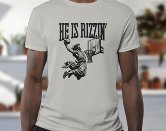 May include: A light gray t-shirt featuring a black graphic of a figure dunking a basketball. The text "HE IS RIZZIN'" is printed above the figure. The shirt is made of a soft material and has a crew neck.