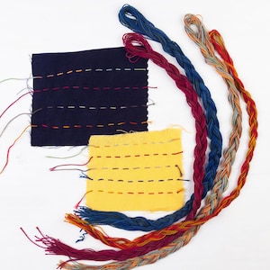 May include: An overhead shot shows two fabric swatches, one navy blue and one yellow, each with colorful hand-stitched lines. Braided embroidery floss in blue, red, and multicolored strands are arranged around the swatches, showcasing textile art.