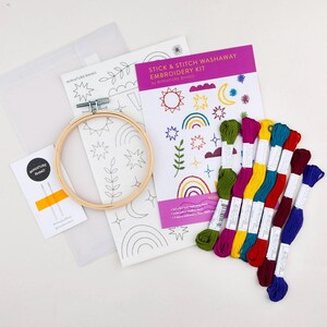 Op de afbeelding: Borduurpakket met een houten ring, naalden en kleurrijk garen. De kit bevat een boekje met een paarse omslag met de tekst "Stick & Stitch Washaway Embroidery Kit". De kit heeft een ontwerp van een regenboog, zon, sterren en andere vormen.