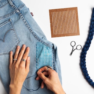 May include: Light blue denim jeans with a blue patch being sewn on. A brown weaving template, small scissors, and a blue braided cord are also present. The jeans have a red Levi's tag.