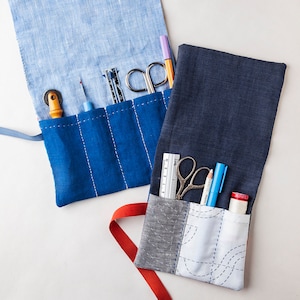 Puede incluir: Dos rollos de herramientas de costura abiertos. Uno es azul claro con un bolsillo azul oscuro, el otro es azul oscuro con un bolsillo gris y blanco. Cada rollo contiene herramientas de costura como tijeras, reglas y bolígrafos. Los rollos están sobre una superficie blanca.