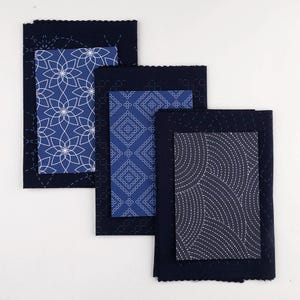 Op de afbeelding: Drie rechthoekige stoffen stukken met ingewikkelde patronen. Elk stuk heeft een donkerblauwe rand met een lichter blauw patroon in het midden. De patronen omvatten bloemen-, geometrische en gebogen lijnen. De randen zijn gestikt met witte draad.