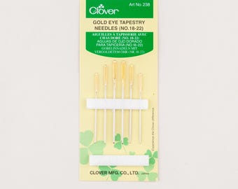 Może przedstawiać: Opakowanie igieł do gobelinów Clover Gold Eye (nr 18-22). Opakowanie jest żółto-zielone z logo Clover i informacjami o produkcie. Igły są złote z srebrnymi trzonkami, utrzymywane przez białą piankę. Widoczny jest napis "GOLD EYE TAPESTRY NEEDLES".