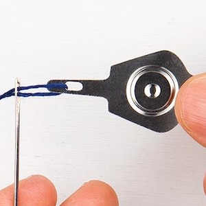 May include: A close-up of a needle being threaded with dark blue thread using a metal needle threader. The threader is held by a person's fingers. The needle is silver and the threader is dark gray.