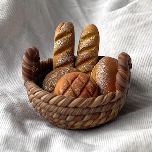 Könnte beinhalten: Ein geflochtener Korb gefüllt mit verschiedenen Brotsorten. Der Korb enthält zwei lange Baguettes, mehrere runde Brötchen und ein Brot mit einem Rautenmuster. Das Brot hat eine goldbraune Farbe, und einige Stücke sind mit weißem Puder bestäubt.