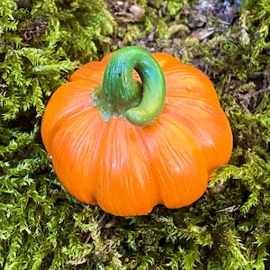 Könnte beinhalten: Eine kleine, runde, orangefarbene Kürbisfigur mit einer glatten, glänzenden Oberfläche, vertikalen Rillen und einem markanten, geschwungenen, hellgrünen Stiel in der Mitte, die eine realistische Form und kontrastreiche Farben aufweist.