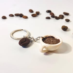 Può includere: Un portachiavi a tema caffè con una tazzina da caffè bianca in miniatura piena di liquido marrone e un ciondolo a forma di chicco di caffè marrone. Il portachiavi ha un anello e una catena argentati. Chicchi di caffè sparsi sullo sfondo.