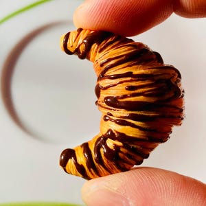 Könnte beinhalten: Ein Miniatur-Croissant mit goldbrauner Außenseite und dunklem Schokoladenüberzug. Das Gebäck wird zwischen zwei Fingern gehalten, was seine geringe Größe und detaillierte Textur zeigt. Die Halbmondform des Croissants ist deutlich definiert, mit sichtbaren Schichten.