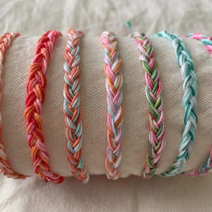 May include: A collection of seven braided friendship bracelets in various pastel colors, including pink, orange, green, and blue. The bracelets are displayed on a white surface, showcasing their woven design and vibrant hues.