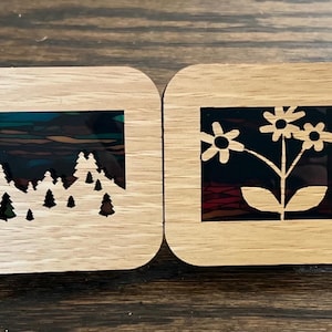 May include: Four wooden square coasters with laser cut designs. Each coaster features a different design: a flower, a mountain range, a flower, and a mountain range. The coasters are made of light brown wood and have a natural finish.