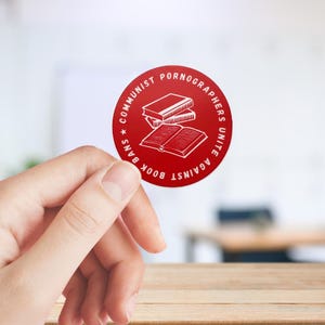 May include: Red round sticker with white text that reads "Communist Pornographers Unite Against Book Bans" and a graphic of a stack of books.