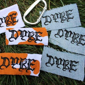 May include: Collection of fabric patches and a lighter on grass. Patches feature the word "DYKE" in black gothic lettering. Some patches are orange and white, others are denim. A silver carabiner clip is also present.