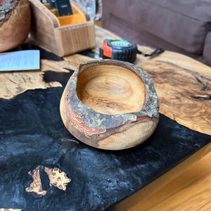 May include: A handcrafted wooden bowl with a natural edge. The bowl is made of light-colored wood with a reddish-brown rim and bark accents. The bowl sits on a black and wood-toned table.