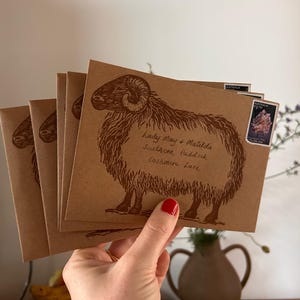 May include: A hand holds a stack of brown envelopes featuring a sheep illustration. The envelopes have text and a postage stamp. The sheep illustration is in brown ink. The envelopes are in front of a vase with flowers.