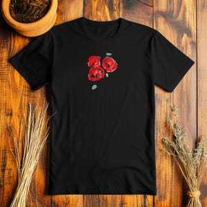 May include: Black t-shirt featuring a floral design of three red poppies with green leaves. The poppies are stylized with detailed petals and a simple stem. The shirt is made of a soft material.