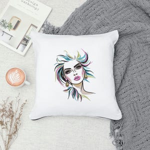Puede incluir: Almohada blanca con una ilustraci&oacute;n colorida de la cara de una mujer con labios rosados y ojos azules. El cabello de la mujer est&aacute; peinado en un mo&ntilde;o desordenado con mechones rosas, azules y amarillos.