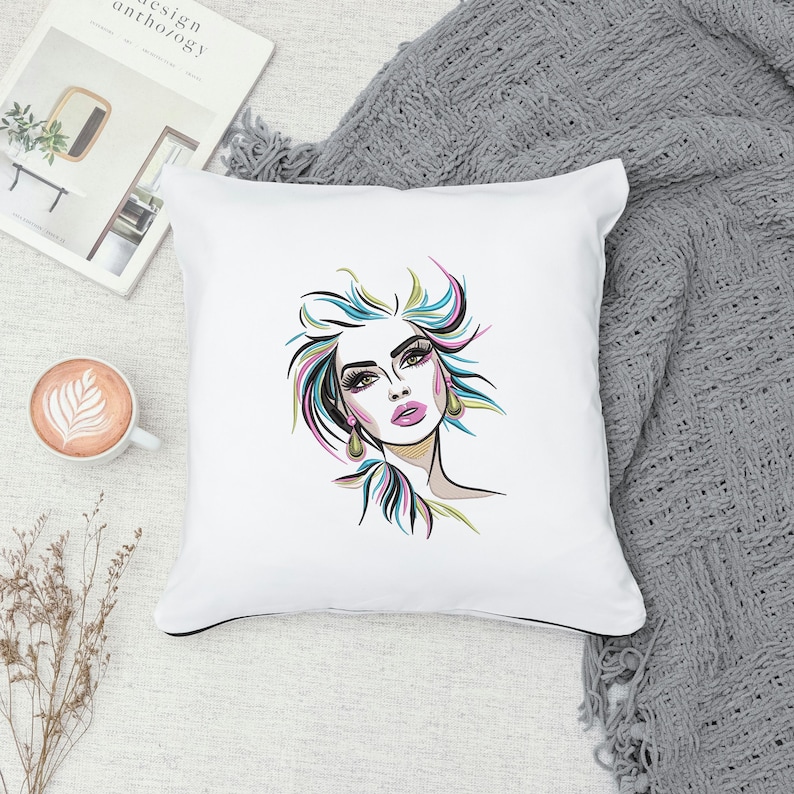 Puede incluir: Almohada blanca con una ilustraci&oacute;n colorida de la cara de una mujer con labios rosados y ojos azules. El cabello de la mujer est&aacute; peinado en un mo&ntilde;o desordenado con mechones rosas, azules y amarillos.