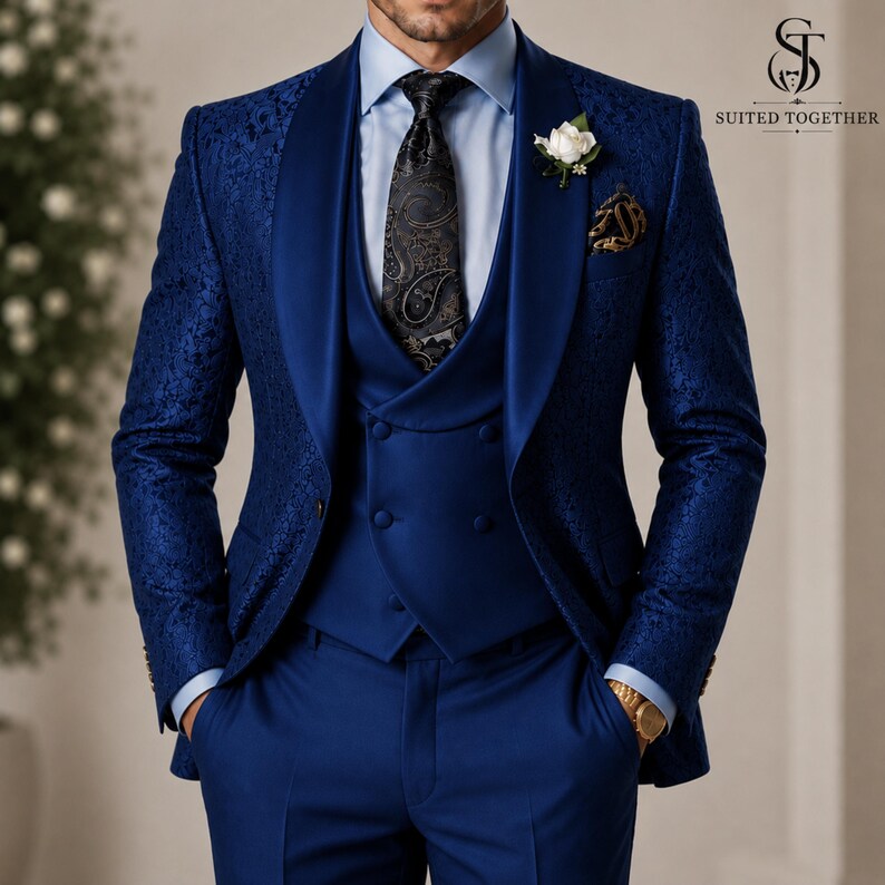 May include: A royal blue three-piece suit featuring a textured floral pattern on the jacket, a solid blue double-breasted vest, a light blue dress shirt, and a black paisley tie. A white boutonniere is pinned to the lapel of the jacket.