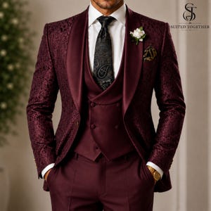 May include: A burgundy three-piece suit featuring a blazer with a tonal floral pattern and a satin shawl lapel, a matching double-breasted vest, a white dress shirt, and a black paisley tie. A white flower boutonniere is pinned to the lapel.