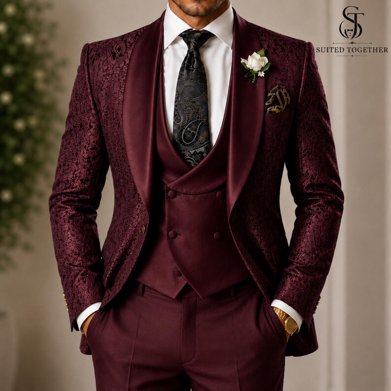 May include: A burgundy three-piece suit featuring a blazer with a tonal floral pattern and a satin shawl lapel, a matching double-breasted vest, a white dress shirt, and a black paisley tie. A white flower boutonniere is pinned to the lapel.