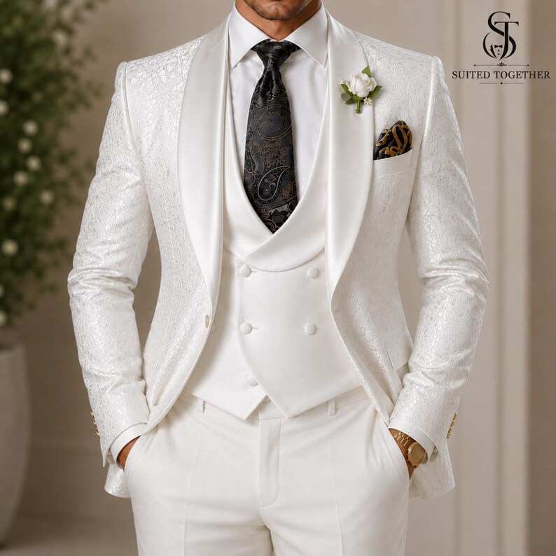 May include: A white three-piece suit featuring a patterned blazer with a shawl lapel, a double-breasted vest, and matching trousers. A black paisley tie is worn with a white shirt, and a white flower boutonniere is pinned to the lapel.