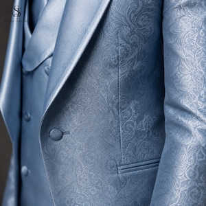 May include: A light blue blazer and matching vest set featuring a tonal damask floral pattern. The blazer has a notched lapel and a single fabric-covered button closure. The vest is double-breasted with fabric-covered buttons and a smooth finish.