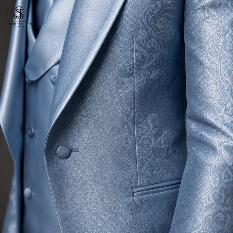 May include: A light blue blazer and matching vest set featuring a tonal damask floral pattern. The blazer has a notched lapel and a single fabric-covered button closure. The vest is double-breasted with fabric-covered buttons and a smooth finish.