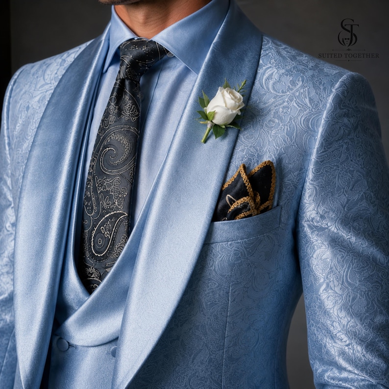 May include: A light blue patterned blazer with a shawl lapel, worn with a matching light blue shirt, a black paisley tie, and a white rose boutonniere. A dark pocket square with gold trim is tucked into the chest pocket. The logo Suited Together is visible.