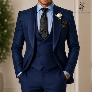May include: A dark blue suit featuring a textured floral pattern on the jacket, a solid blue double-breasted vest, a light blue dress shirt, and a dark patterned tie. A white flower boutonniere is pinned to the lapel, and a gold watch is visible.