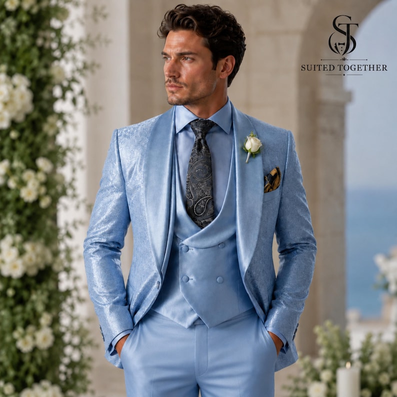 May include: A light blue suit featuring a patterned blazer, a double-breasted waistcoat, and matching trousers. The ensemble includes a light blue dress shirt, a dark grey paisley tie, and a white floral boutonniere on the lapel.