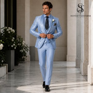 May include: A light blue three-piece suit featuring a patterned blazer, matching vest, and trousers. The ensemble includes a dark patterned tie, a white dress shirt, and a white floral boutonniere. The logo Suited Together is visible.