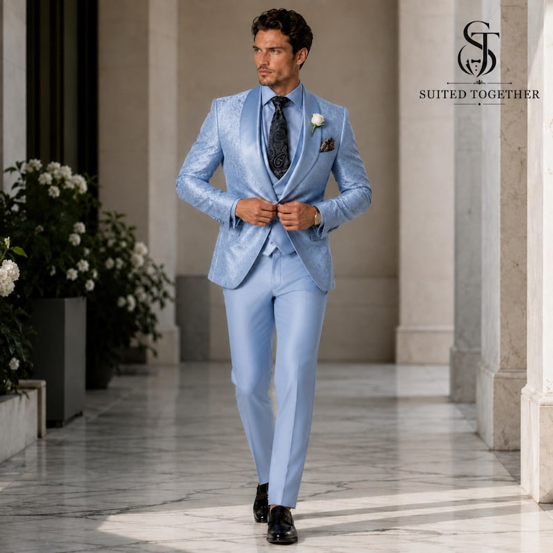 May include: A light blue three-piece suit featuring a patterned blazer, matching vest, and trousers. The ensemble includes a dark patterned tie, a white dress shirt, and a white floral boutonniere. The logo Suited Together is visible.