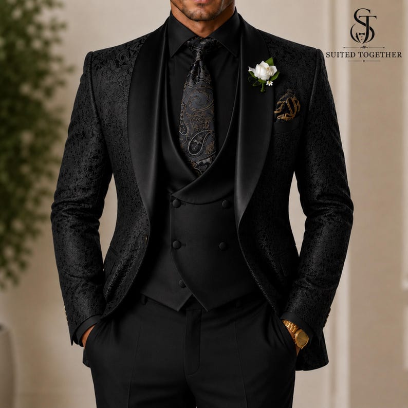 May include: A black three-piece suit featuring a patterned blazer with a satin shawl lapel, a solid black double-breasted vest, and a black paisley tie. A white floral boutonniere is pinned to the lapel. The logo SUITED TOGETHER is visible.