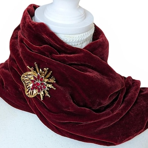 Op de afbeelding: Bordeaux fluwelen sjaal gedrapeerd over een witte mannequin. Een goudkleurige broche met rode edelstenen is op de sjaal gespeld. De sjaal heeft een zachte textuur en is afgewerkt met kwastjes.