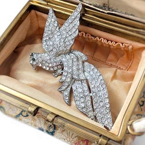 Puede incluir: Un broche vintage plateado con forma de pájaro, incrustado con pedrería transparente, descansa dentro de un pequeño bolso adornado con marco dorado. El bolso tiene un estampado floral en el exterior y un forro color melocotón. El asa del bolso es visible.