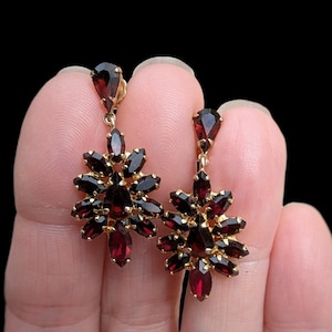 May include: A pair of vintage gold-tone earrings with a floral design. Each earring features a teardrop-shaped stone at the top and a cluster of dark red, oval-shaped stones forming a flower. The earrings are set against a black background.