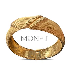 May include: A gold-toned bracelet with a textured, ridged design. The bracelet has a hinged clasp and the word "MONET" is printed on the front. The bracelet is set against a white background.