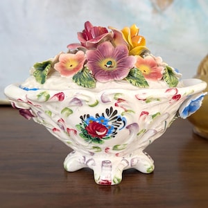 Op de afbeelding: Een decoratieve, witte keramische sieradenschaal met deksel, versierd met kleurrijke, handgeschilderde bloemmotieven. Het deksel is bekroond met een cluster van gebeeldhouwde bloemen in roze, geel en paars. De schaal heeft een geschulpte rand en een voet.