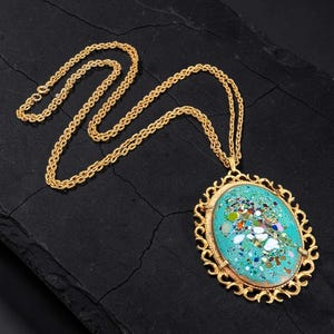 May include: A gold-tone necklace featuring a long chain and an oval pendant. The pendant has an ornate, filigree-style gold frame surrounding a central turquoise-colored inlay speckled with multicolored, reflective metallic flecks and shards.