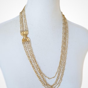 May include: A gold-coloured necklace with multiple strands of fine chain. The necklace has a decorative clasp with a floral design. The chains hang down, creating a layered effect. The necklace is displayed on a white mannequin.