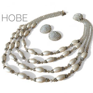 Puede incluir: Un conjunto de joyas vintage con un collar de múltiples hilos y pendientes a juego. El collar tiene cuentas ovaladas de color blanco roto con detalles de cristal, y los pendientes son redondos, incrustados con cristales brillantes. La palabra "HOBE" es visible.
