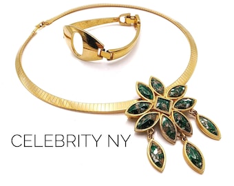 May include: A gold necklace with a green stone pendant in the shape of a flower. The pendant has a square center and is surrounded by eight petals. The necklace is on a white background.