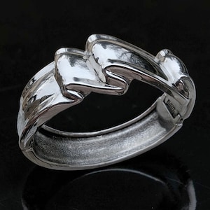 May include: A silver-coloured hinged bangle bracelet with a flowing, sculptural design. The bracelet features interconnected, wave-like forms, creating a dynamic and modern aesthetic. The metal has a polished finish, reflecting light and adding to its visual appeal.