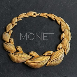 Könnte beinhalten: Eine goldfarbene Halskette mit Blattdesign. Die Halskette hat eine Reihe von detaillierten, strukturierten Blättern, die miteinander verbunden sind. Das Wort "MONET" ist auf dem schwarzen Hintergrund aufgedruckt. Ein auffälliges Schmuckstück.