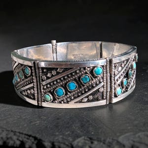 Può includere: Bracciale color argento con pietre turchesi. Il bracciale presenta un design intricato con motivi geometrici e piccoli accenti rotondi turchesi. La chiusura è visibile nella parte superiore. Il bracciale sembra essere in metallo.