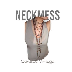 May include: A layered necklace set with multiple chains and beads, including a silver pendant and a large round charm. The necklace is worn over a light gray tank top. The text "Neckmess Curated Vintage" is visible at the bottom of the image.